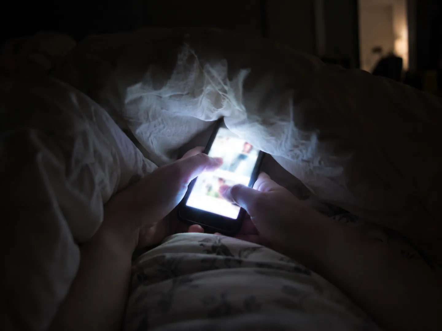 Hands holding a glowing smartphone under the covers in a dark bedroom, the screen light falling on the duvet